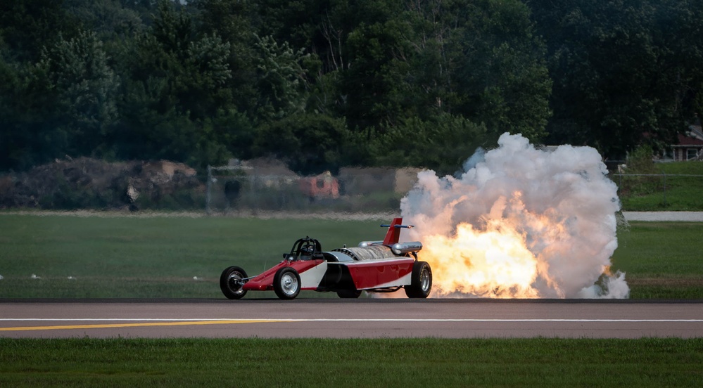 A red and white jet-powered dragster or rocket car is traveling along what appears to be an airstrip or runway, producing a massive plume of fire and white smoke from its rear-mounted jet/rocket engine. The vehicle appears to be in the middle of a performance run, likely at an airshow or motorsport event. The dramatic exhaust flame and smoke cloud dominate the right side of the image.