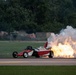 A red and white jet-powered dragster or rocket car is traveling along what appears to be an airstrip or runway, producing a massive plume of fire and white smoke from its rear-mounted jet/rocket engine. The vehicle appears to be in the middle of a performance run, likely at an airshow or motorsport event. The dramatic exhaust flame and smoke cloud dominate the right side of the image.