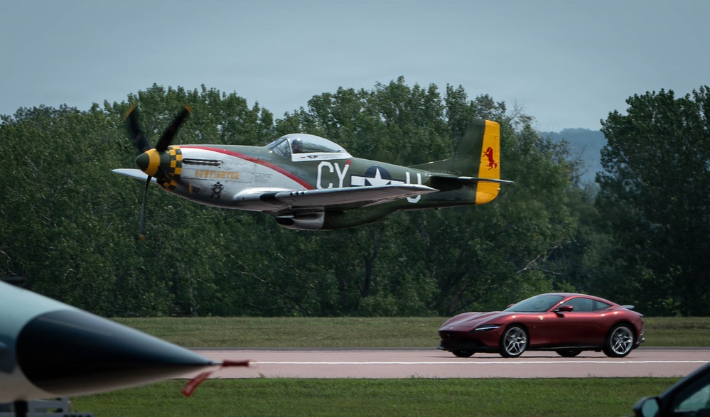 A WWII-era North American P-51 Mustang fighter aircraft flies at very low altitude over an airfield runway during what appears to be an airshow. The aircraft bears the markings 'CY-H' and features a yellow tail with a red horse emblem, along with a checkered yellow-and-black nose cowling. A modern red Ferrari Roma sports car is visible on or near the runway in the background, suggesting a staged speed comparison event.