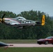 A WWII-era North American P-51 Mustang fighter aircraft flies at very low altitude over an airfield runway during what appears to be an airshow. The aircraft bears the markings 'CY-H' and features a yellow tail with a red horse emblem, along with a checkered yellow-and-black nose cowling. A modern red Ferrari Roma sports car is visible on or near the runway in the background, suggesting a staged speed comparison event.