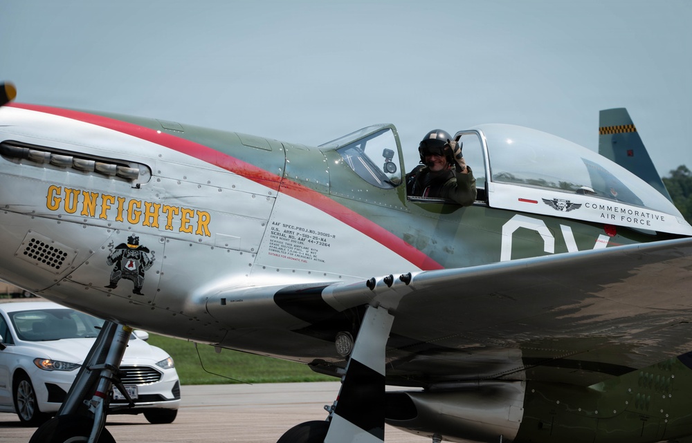 A restored North American P-51 Mustang named 'Gunfighter' sits on a tarmac, operated by the Commemorative Air Force. A pilot wearing a flight helmet is visible in the open cockpit, appearing to wave or give a thumbs-up gesture. The aircraft features a polished aluminum finish with a red stripe and period-accurate nose art.