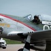 A restored North American P-51 Mustang named 'Gunfighter' sits on a tarmac, operated by the Commemorative Air Force. A pilot wearing a flight helmet is visible in the open cockpit, appearing to wave or give a thumbs-up gesture. The aircraft features a polished aluminum finish with a red stripe and period-accurate nose art.