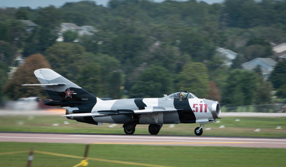 A Soviet-era MiG-17 (or Chinese J-5 variant) jet fighter painted in a black-and-white camouflage scheme with the number '511' in red on the fuselage is taxiing or taking off on a runway. The aircraft displays a red star insignia on the tail fin and bears the FAA registration 'N6963X', indicating it is a privately operated warbird in the United States. The background shows motion blur consistent with a panning camera technique, with trees and residential structures visible beyond the airfield.