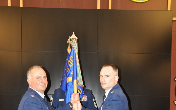 Two U.S. Air Force officers in service dress uniforms are participating in a formal guidon/flag transfer ceremony, likely a change of command. One officer on the left is passing a blue unit guidon to the officer on the right, both gripping the staff simultaneously. A third uniformed individual stands at attention in the background holding the flag staff position.
