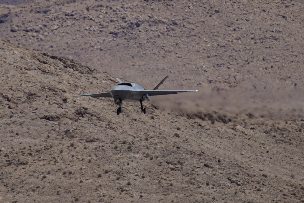 A gray military aircraft, appearing to be a stealth-configured jet or advanced drone, is captured at very low altitude over arid desert terrain. The aircraft has its landing gear deployed and is kicking up a dust cloud behind it, suggesting it is either taking off or landing on an unprepared surface. The rocky, scrub-covered hillside in the background is consistent with a desert test range environment.