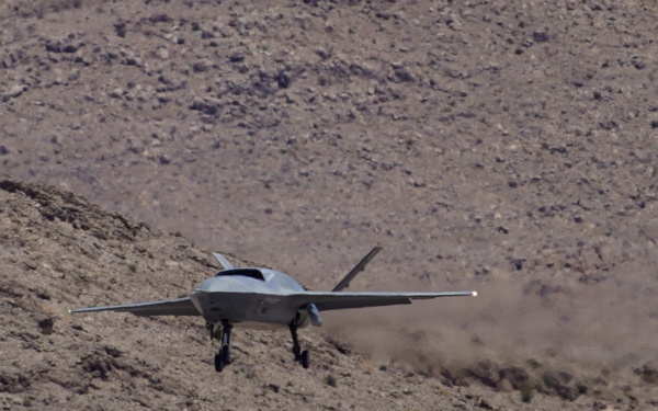 A gray military aircraft, appearing to be a stealth-configured jet or advanced drone, is captured at very low altitude over arid desert terrain. The aircraft has its landing gear deployed and is kicking up a dust cloud behind it, suggesting it is either taking off or landing on an unprepared surface. The rocky, scrub-covered hillside in the background is consistent with a desert test range environment.