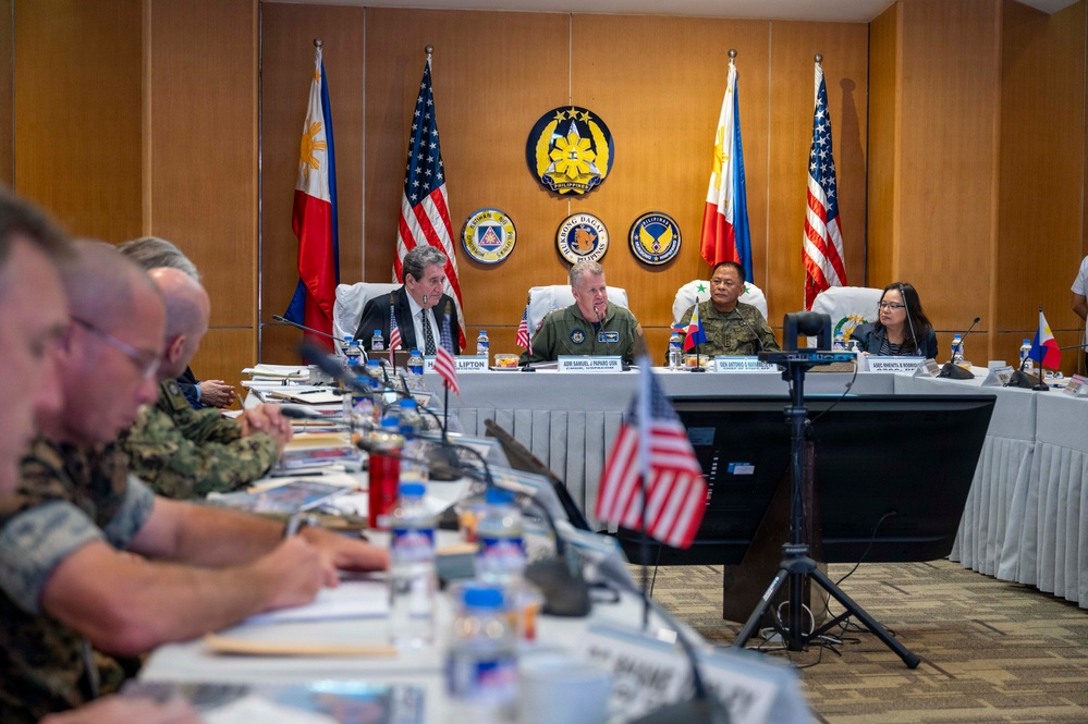 A formal bilateral military meeting between United States and Philippine armed forces officials is taking place in a conference room. Senior military officers and civilian officials are seated at a head table with name placards, flanked by Philippine and American flags, beneath an official Philippine military seal. Additional military personnel are seated along a side table, taking notes.