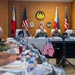 A formal bilateral military meeting between United States and Philippine armed forces officials is taking place in a conference room. Senior military officers and civilian officials are seated at a head table with name placards, flanked by Philippine and American flags, beneath an official Philippine military seal. Additional military personnel are seated along a side table, taking notes.