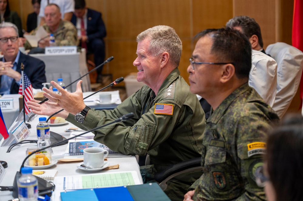 A formal military meeting or conference is taking place at a large table, featuring personnel in U.S. and what appears to be Philippine military uniforms. A senior U.S. military officer in an olive green flight suit with an American flag patch and three-star insignia is speaking and gesturing, seated next to an Asian military officer in camouflage uniform. The setting includes name placards, microphones, water bottles, and small national flags, indicating a bilateral or multilateral military dialogue.