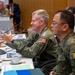 A formal military meeting or conference is taking place at a large table, featuring personnel in U.S. and what appears to be Philippine military uniforms. A senior U.S. military officer in an olive green flight suit with an American flag patch and three-star insignia is speaking and gesturing, seated next to an Asian military officer in camouflage uniform. The setting includes name placards, microphones, water bottles, and small national flags, indicating a bilateral or multilateral military dialogue.