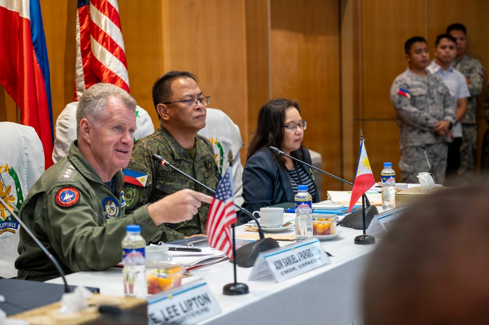 A formal military and diplomatic meeting between U.S. and Philippine officials, likely a bilateral defense consultation. A U.S. military officer in flight suit is speaking and gesturing, seated alongside Philippine military personnel and a civilian official. Name placards, small national flags, and formal table settings indicate an official high-level conference.