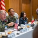A formal military and diplomatic meeting between U.S. and Philippine officials, likely a bilateral defense consultation. A U.S. military officer in flight suit is speaking and gesturing, seated alongside Philippine military personnel and a civilian official. Name placards, small national flags, and formal table settings indicate an official high-level conference.