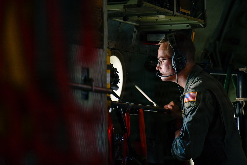 A U.S. military airman is seated inside what appears to be a military cargo or transport aircraft, wearing a flight suit with an American flag patch and a communications headset. He is focused on reviewing documents or a clipboard in a dimly lit interior environment. The setting suggests an active flight operation or mission briefing aboard the aircraft.
