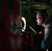 A U.S. military airman is seated inside what appears to be a military cargo or transport aircraft, wearing a flight suit with an American flag patch and a communications headset. He is focused on reviewing documents or a clipboard in a dimly lit interior environment. The setting suggests an active flight operation or mission briefing aboard the aircraft.