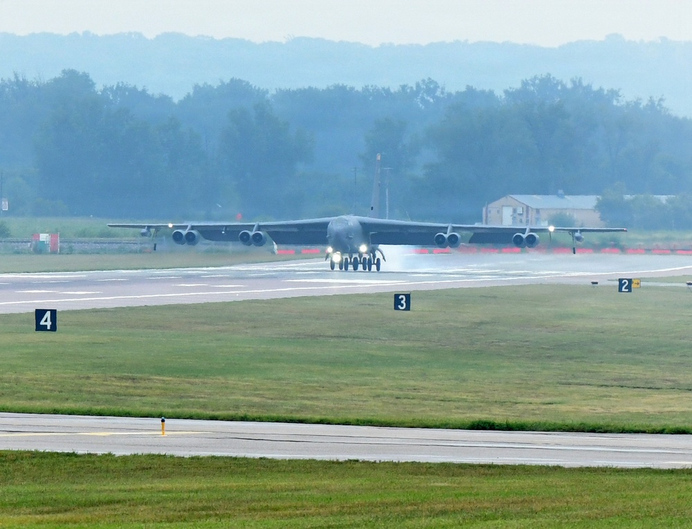 A large multi-engine bomber aircraft, consistent with a B-52 Stratofortress, is taxiing or rolling on a wet runway at a military airfield. The runway surface appears wet, likely from recent rain, with visible spray or mist around the landing gear. Distance markers numbered 2, 3, and 4 are visible along the runway edge.