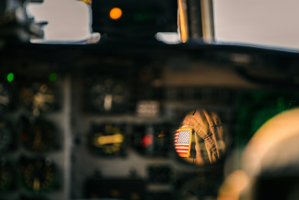 The image shows a cockpit interior of a military aircraft, with a small circular mirror reflecting a crew member's arm bearing a U.S. flag patch. The cockpit instruments and panels are visible but largely out of focus in the background. The composition deliberately focuses on the mirror reflection to highlight the American flag insignia on the flight suit.