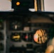 The image shows a cockpit interior of a military aircraft, with a small circular mirror reflecting a crew member's arm bearing a U.S. flag patch. The cockpit instruments and panels are visible but largely out of focus in the background. The composition deliberately focuses on the mirror reflection to highlight the American flag insignia on the flight suit.