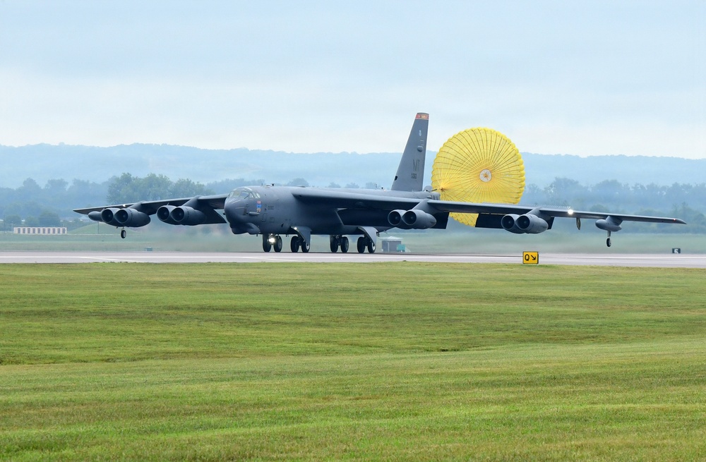 A B-52 Stratofortress strategic bomber is landing on a wet runway with its yellow drag parachute fully deployed to assist in deceleration. The aircraft is painted in dark gray and features eight engines mounted in four underwing pods. The scene appears to be at a military airfield with green grass, overcast skies, and tree-covered hills visible in the background.
