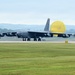 A B-52 Stratofortress strategic bomber is landing on a wet runway with its yellow drag parachute fully deployed to assist in deceleration. The aircraft is painted in dark gray and features eight engines mounted in four underwing pods. The scene appears to be at a military airfield with green grass, overcast skies, and tree-covered hills visible in the background.