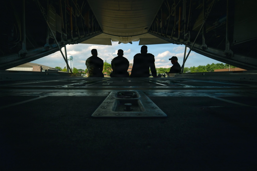 The image is taken from inside a military cargo aircraft looking outward through the open rear ramp. Four individuals are silhouetted against a bright sky, seated or standing at the edge of the ramp. A military airfield with buildings and a control tower is visible in the background.
