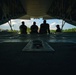 The image is taken from inside a military cargo aircraft looking outward through the open rear ramp. Four individuals are silhouetted against a bright sky, seated or standing at the edge of the ramp. A military airfield with buildings and a control tower is visible in the background.
