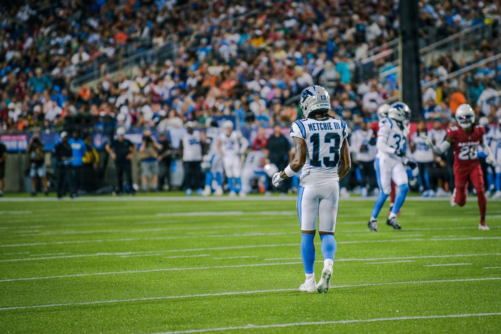 An NFL football game is in progress featuring a player wearing jersey number 13 with the name 'METCHIE III' on the back, dressed in white and light blue Carolina Panthers-style uniforms. The opposing team appears to be the Arizona Cardinals, identifiable by a player in a red jersey with number 28. The game is being played in a large stadium filled with spectators.