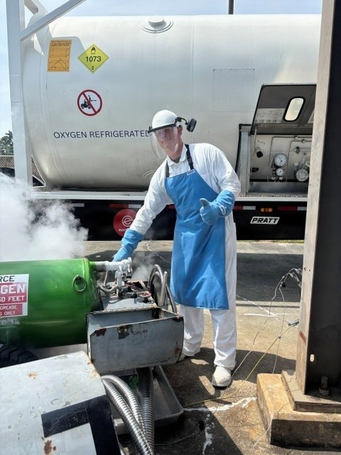 A worker in full personal protective equipment is handling a cryogenic oxygen transfer operation outdoors. Visible vapor/fog indicates the presence of liquid oxygen (LOX) being transferred from a tanker truck labeled 'OXYGEN REFRIGERATED' with UN hazmat placard 1073. The worker is giving a thumbs-up gesture while managing the transfer connections.