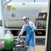 A worker in full personal protective equipment is handling a cryogenic oxygen transfer operation outdoors. Visible vapor/fog indicates the presence of liquid oxygen (LOX) being transferred from a tanker truck labeled 'OXYGEN REFRIGERATED' with UN hazmat placard 1073. The worker is giving a thumbs-up gesture while managing the transfer connections.