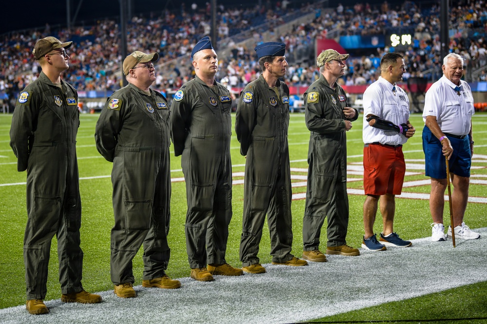 A group of military personnel in olive green flight suits stand at attention on an NFL football field during what appears to be a pre-game or halftime ceremony. They are joined by two civilians, one of whom is using a walking cane. The event is taking place at a well-attended night game in a large stadium.