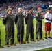 A group of military personnel in olive green flight suits stand at attention on an NFL football field during what appears to be a pre-game or halftime ceremony. They are joined by two civilians, one of whom is using a walking cane. The event is taking place at a well-attended night game in a large stadium.