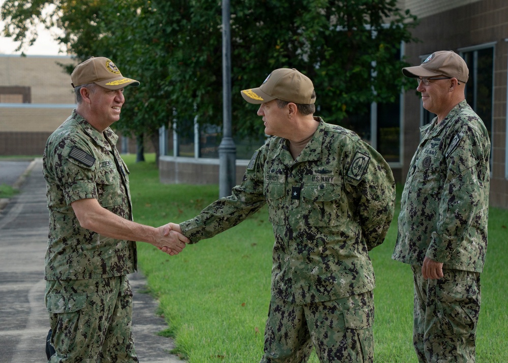 Three U.S. Navy personnel in NWU Type III (woodland digital camouflage) uniforms are gathered outdoors on a military installation. Two of the individuals are shaking hands in what appears to be a formal greeting or recognition ceremony, while a third stands nearby observing. All three wear tan/coyote brown ball caps with command insignia patches.