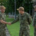 Three U.S. Navy personnel in NWU Type III (woodland digital camouflage) uniforms are gathered outdoors on a military installation. Two of the individuals are shaking hands in what appears to be a formal greeting or recognition ceremony, while a third stands nearby observing. All three wear tan/coyote brown ball caps with command insignia patches.