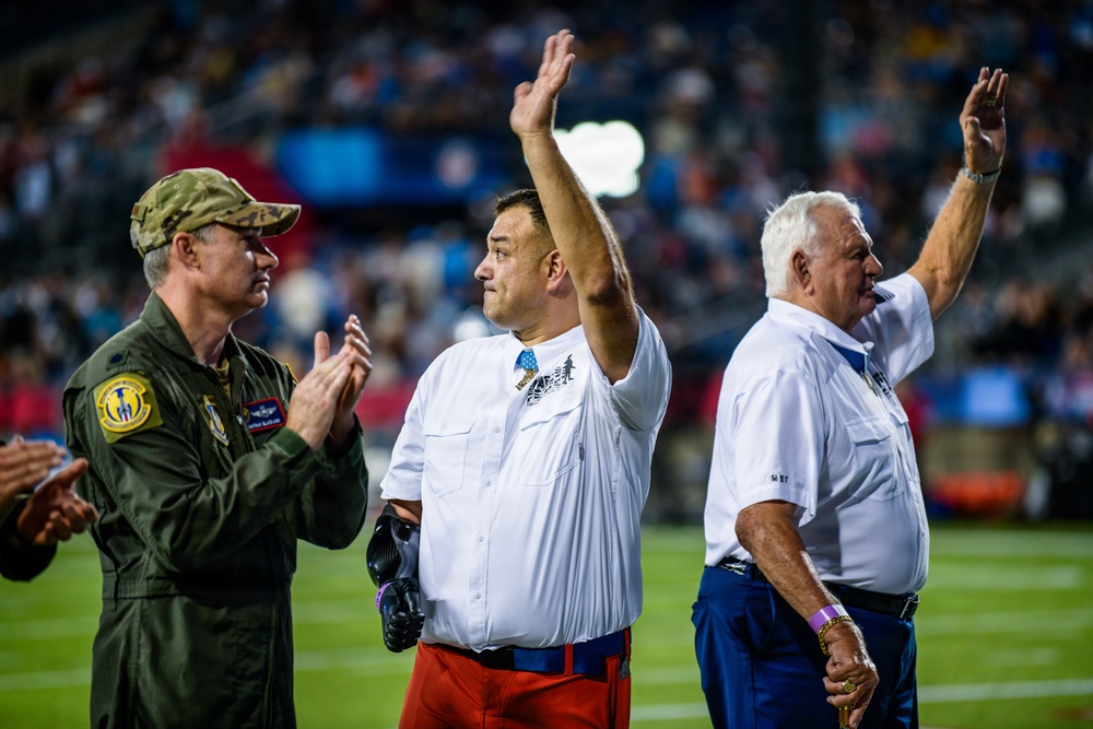 Three men stand on what appears to be a football field during a ceremony, with two of them raising their hands to acknowledge a crowd. One man is wearing a military flight suit with patches, while the other two are dressed in white shirts. The stadium is filled with spectators in the background, suggesting a public recognition or honorary event.