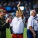 Three men stand on what appears to be a football field during a ceremony, with two of them raising their hands to acknowledge a crowd. One man is wearing a military flight suit with patches, while the other two are dressed in white shirts. The stadium is filled with spectators in the background, suggesting a public recognition or honorary event.