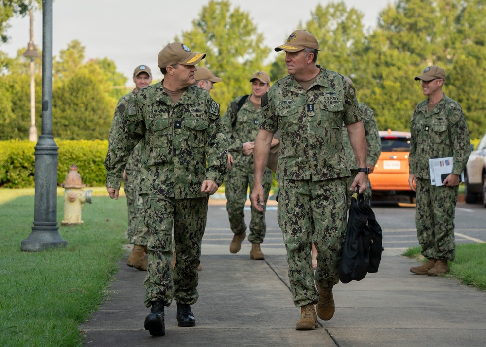 A group of U.S. Navy personnel in NWU Type III (green camouflage) uniforms are walking along a sidewalk at what appears to be a military installation. Two senior officers are in the foreground engaged in conversation, with additional personnel following behind. The setting appears to be a well-maintained base with green landscaping and parked vehicles visible in the background.