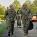 A group of U.S. Navy personnel in NWU Type III (green camouflage) uniforms are walking along a sidewalk at what appears to be a military installation. Two senior officers are in the foreground engaged in conversation, with additional personnel following behind. The setting appears to be a well-maintained base with green landscaping and parked vehicles visible in the background.