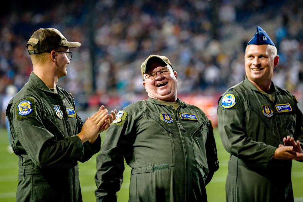 Three individuals wearing olive green military flight suits are standing on what appears to be a sports stadium field during a nighttime event. Two of the men are wearing camouflage baseball caps while the third wears a blue military garrison cap. They appear to be laughing and clapping, suggesting they are being recognized or honored at a public event.