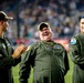Three individuals wearing olive green military flight suits are standing on what appears to be a sports stadium field during a nighttime event. Two of the men are wearing camouflage baseball caps while the third wears a blue military garrison cap. They appear to be laughing and clapping, suggesting they are being recognized or honored at a public event.