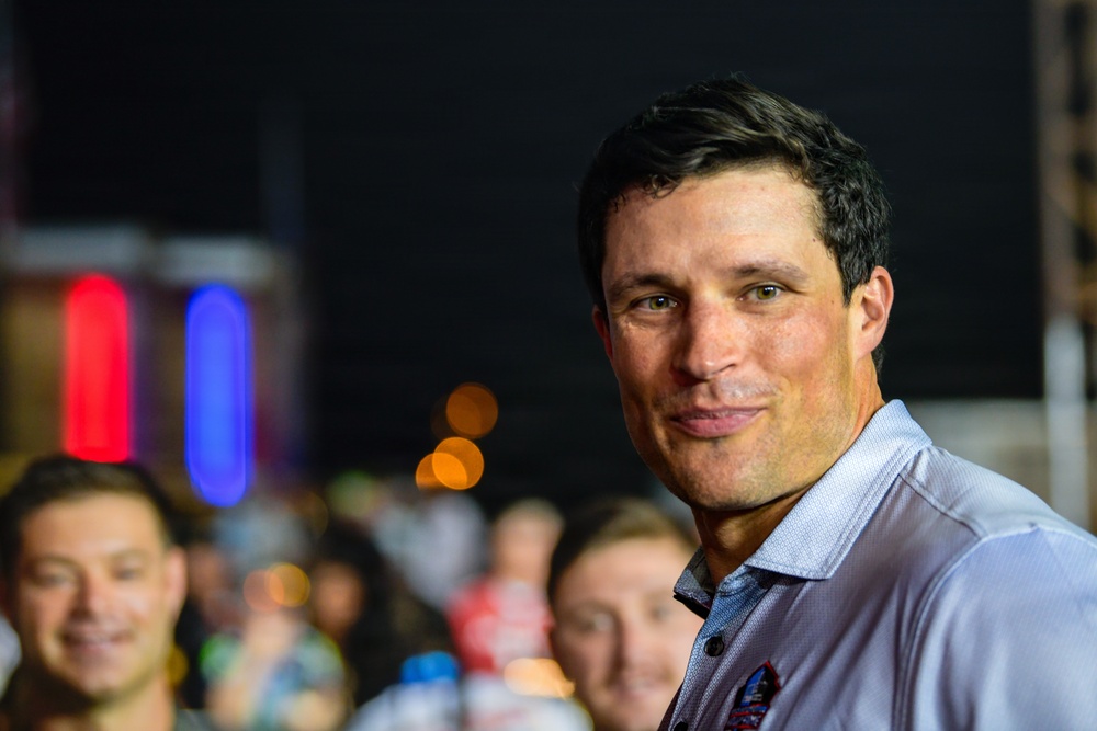 A man with dark hair is the primary subject of this nighttime photograph, appearing to be at an outdoor event or gathering. He is wearing a light blue collared shirt with what appears to be a small logo or pin on it. The background shows a crowd of people and colorful bokeh lights in red, blue, and orange.