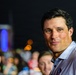 A man with dark hair is the primary subject of this nighttime photograph, appearing to be at an outdoor event or gathering. He is wearing a light blue collared shirt with what appears to be a small logo or pin on it. The background shows a crowd of people and colorful bokeh lights in red, blue, and orange.