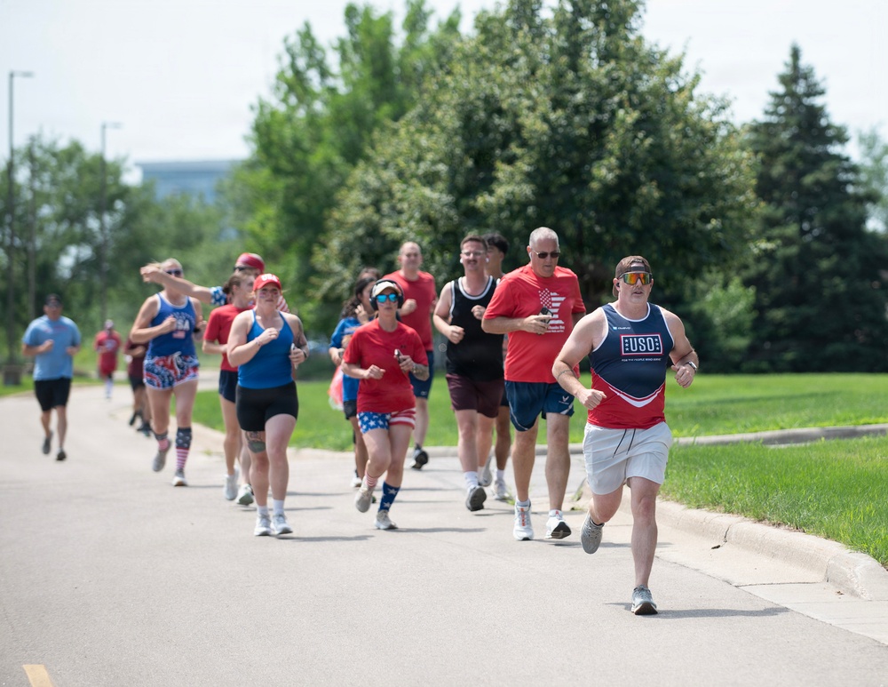 A group of runners participates in what appears to be a patriotic-themed fun run or charity race, with many participants wearing red, white, and blue attire. The lead runner wears a USO-branded tank top, suggesting this may be a military-affiliated or USO-sponsored event. The race takes place on a paved path surrounded by green grass and trees on a sunny day.