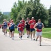 A group of runners participates in what appears to be a patriotic-themed fun run or charity race, with many participants wearing red, white, and blue attire. The lead runner wears a USO-branded tank top, suggesting this may be a military-affiliated or USO-sponsored event. The race takes place on a paved path surrounded by green grass and trees on a sunny day.