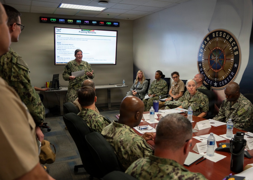 A military briefing is taking place at the Navy Human Resources Service Center, with a female Navy officer presenting 'Framing Remarks' on a projected screen to a group of uniformed personnel seated around a conference table. The attendees are primarily wearing Navy Working Uniform (NWU) camouflage pattern, with at least one civilian present. The setting appears to be a formal command briefing room equipped with multiple time zone clocks on the wall.