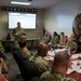 A military briefing is taking place at the Navy Human Resources Service Center, with a female Navy officer presenting 'Framing Remarks' on a projected screen to a group of uniformed personnel seated around a conference table. The attendees are primarily wearing Navy Working Uniform (NWU) camouflage pattern, with at least one civilian present. The setting appears to be a formal command briefing room equipped with multiple time zone clocks on the wall.