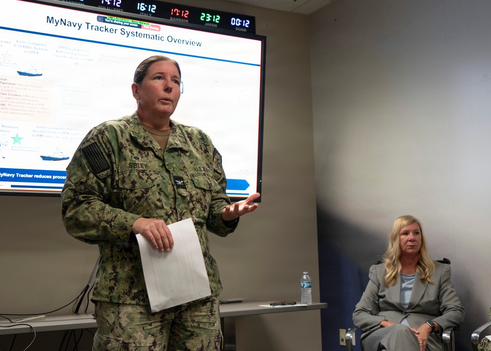 A U.S. Navy officer identified by name tape as 'SIBLEY' is presenting a briefing on the 'MyNavy Tracker Systematic Overview' displayed on a large screen behind her. She is holding papers and gesturing while speaking, with a civilian woman in a gray blazer seated to the right. A digital world clock on the wall displays times for multiple locations including Italy, Bahrain, Japan, and Guam.