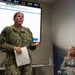A U.S. Navy officer identified by name tape as 'SIBLEY' is presenting a briefing on the 'MyNavy Tracker Systematic Overview' displayed on a large screen behind her. She is holding papers and gesturing while speaking, with a civilian woman in a gray blazer seated to the right. A digital world clock on the wall displays times for multiple locations including Italy, Bahrain, Japan, and Guam.