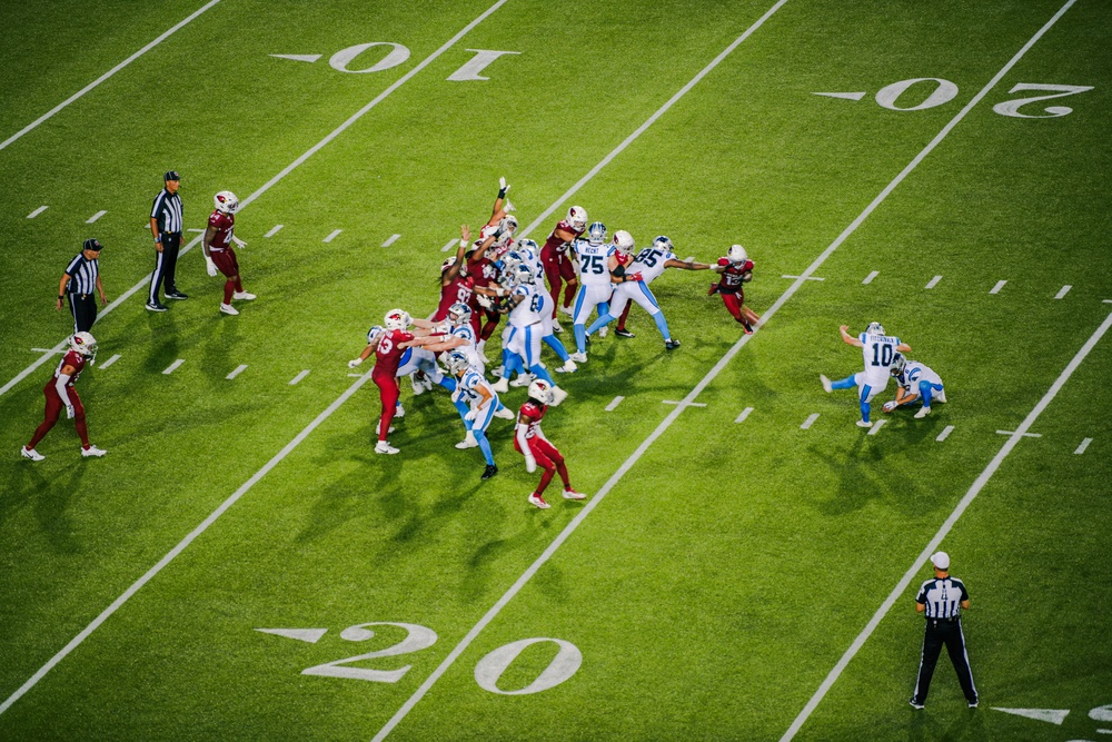 An aerial view of an NFL football game in progress, showing a play developing near the 20-yard line. Two teams are engaged at the line of scrimmage — one in dark red/cardinal uniforms and one in white and blue uniforms. Two officials are visible on the field monitoring the play.