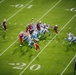 An aerial view of an NFL football game in progress, showing a play developing near the 20-yard line. Two teams are engaged at the line of scrimmage — one in dark red/cardinal uniforms and one in white and blue uniforms. Two officials are visible on the field monitoring the play.
