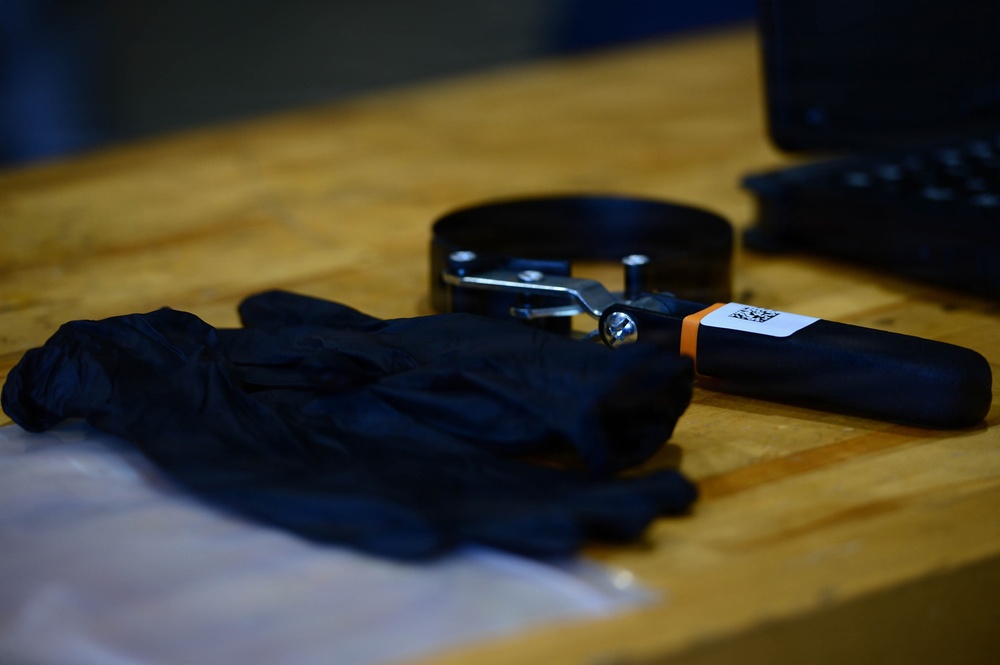 The image shows a wooden workbench surface with mechanical tools and protective equipment laid out. A black nitrile glove, an oil filter wrench with a handle, and what appears to be a filter removal tool are the primary items visible. The setting suggests an automotive maintenance or mechanical work environment.