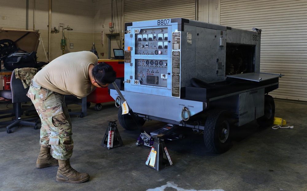 A military service member in MultiCam/OCP camouflage uniform is performing maintenance on a piece of ground support equipment labeled 'B807' inside a maintenance hangar or shop. The equipment appears to be a mobile generator or power unit mounted on a wheeled trailer, with jack stands placed underneath for support. The setting is an indoor military maintenance facility with tools and equipment visible in the background.